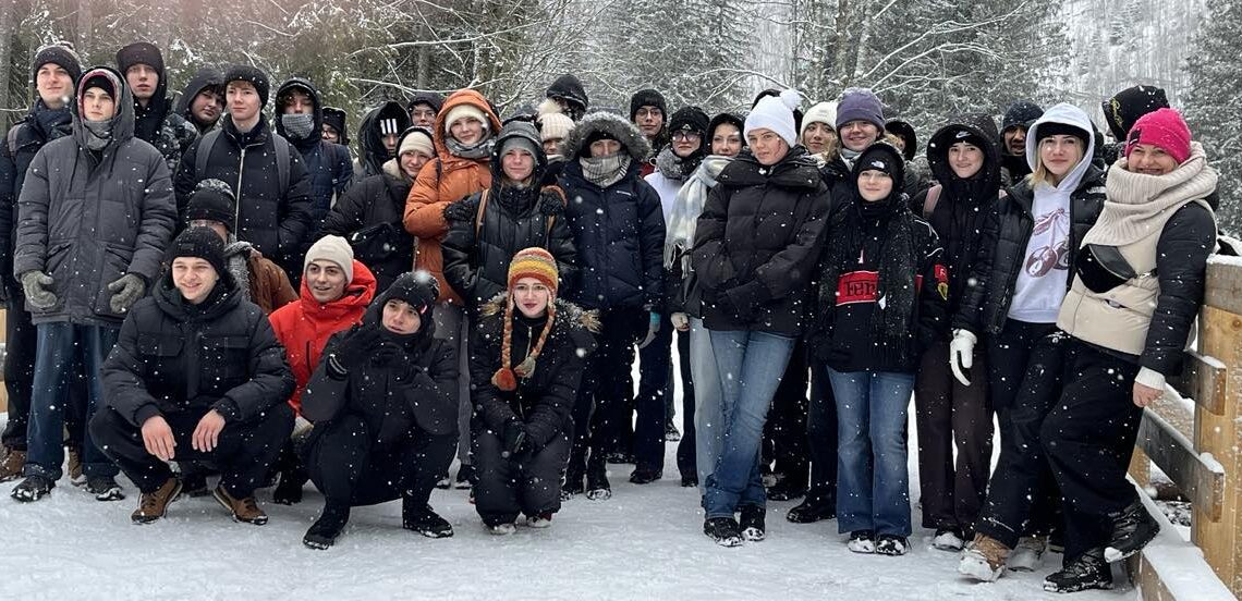 zdjęcie grupowe uczestników wycieczki. Młodzież ubrana w zimowe kurtki, kilkoro z nich w pierwszym rzędzie kuca. Wszyscy są uśmiechnięci i zadowoleni. W tle ośnieżone drzewa.