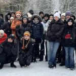 zdjęcie grupowe uczestników wycieczki. Młodzież ubrana w zimowe kurtki, kilkoro z nich w pierwszym rzędzie kuca. Wszyscy są uśmiechnięci i zadowoleni. W tle ośnieżone drzewa.