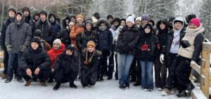 zdjęcie grupowe uczestników wycieczki. Młodzież ubrana w zimowe kurtki, kilkoro z nich w pierwszym rzędzie kuca. Wszyscy są uśmiechnięci i zadowoleni. W tle ośnieżone drzewa. 