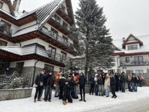 Zdjęcie przedstawia grupę  uczniów, stojących na śniegu przed dużym, drewniano-murowanym budynkiem w stylu zakopiańskim. Budynek ma spadzisty dach pokryty śniegiem, balkony z metalowymi barierkami i jest otoczony zimową scenerią. Po prawej stronie widoczna jest choinka, a wokół pada śnieg. Uczestnicy wycieczki są ubrani w zimowe kurtki, czapki i szaliki, co wskazuje na zimową porę roku. Atmosfera jest radosna, a uczniowie wydają się być zadowoleni.