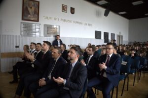 zdjęcie gości wsłuchanych w prelekcję, w tle jasna ściana w tablicami pamiątkowymi