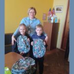 Na zdjęciu znajduje się starsza kobieta z krótkimi blond włosami i okularami, uśmiechająca się delikatnie. Kobieta nosi jasnoniebieską bluzkę z kołnierzykiem oraz identyfikator na smyczy z widocznym zdjęciem, co sugeruje, że może być nauczycielką lub pracownikiem szkoły. Obejmuje ona dwójkę dzieci – dziewczynkę i chłopca – którzy stoją przed nią, trzymając plecaki z motywem roślinnym. Plecaki są w odcieniach zieleni, z wzorem przedstawiającym liście i małe zwierzątka, takie jak lisy i ptaki. Dziewczynka po lewej stronie ma jasną cerę, blond włosy związane w kucyk oraz czarną bluzę. Uśmiecha się delikatnie, trzymając plecak obiema rękami. Chłopiec po prawej stronie również ma jasną cerę i jasne włosy, nosi ciemnoniebieski sweter z jasnym wzorem na wysokości klatki piersiowej. Również trzyma plecak obiema rękami i patrzy na aparat z lekkim uśmiechem. W tle znajduje się żółta ściana, drewniana szafka, na której ustawione są dwie amerykańskie flagi, fotografia przedstawiająca budynek, biała figurka oraz kilka przedmiotów takich jak butelki i dokumenty. Po lewej stronie widoczna jest część ciemnej kanapy. W dolnej części zdjęcia, na stole, leży jeszcze jeden plecak o identycznym wzorze. Całość sceny sprawia wrażenie przyjaznej i pełnej ciepła atmosfery, co może sugerować akcję charytatywną lub przekazanie upominków uczniom.