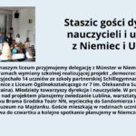 Grafika składa się z dwóch części: zdjęcia po lewej stronie oraz tekstu po prawej stronie na jasnoniebieskim tle. Zdjęcie: Przedstawia scenę w sali szkolnej z drewnianą podłogą i białymi ścianami. Na ścianie w tle widoczny jest ekran z projektorem, na którym wyświetlono obraz przedstawiający krajobraz z wodą i górami. Na scenie stoi siedem osób – pięć dorosłych i dwie młodsze osoby, prawdopodobnie uczniowie. Dorośli są elegancko ubrani – mężczyzna w garniturze, kobiety w swetrach lub marynarkach. Wśród nich jedna osoba trzyma mikrofon, a reszta uśmiecha się i patrzy na publiczność. Publiczność składa się z uczniów siedzących na krzesłach; niektórzy mają widoczne kolorowe ubrania, w tym dziewczyna z długimi rudymi włosami w czerwonej bluzie. Tekst: Po prawej stronie znajduje się blok tekstu w języku polskim. Nagłówek w dużej czarnej czcionce głosi: „Staszic gości dyrekcję, nauczycieli i uczniów z Niemiec i Ukrainy” Poniżej znajduje się akapit tekstu w mniejszej czcionce: Od kilku dni w naszym liceum przyjmujemy delegację z Münster w Niemczech i Winnicy w Ukrainie w ramach wymiany szkolnej realizującej projekt „democracy and media”. Do Lublina przyjechało 14 uczniów ze szkoły partnerskiej Schillegymnasium Münster oraz dwie uczennice z Liceum Ogólnokształcącego nr 7 im. Oleksandra Suchomowskiego w Winnicy (Ukraina). Młodzieży towarzyszy dyrekcja i nauczyciele. W programie oprócz wspólnej pracy nad projektem planujemy zwiedzanie Lublina, warsztaty w Domu Słów, wizytę w Ośrodku Brama Grodzka Teatr NN, wycieczkę do Sandomierza i pobyt studyjny w Państwowym Muzeum na Majdanku. Goście mieszkają w rodzinach uczniów z klasy 2a i 3b. Wymiana potrwa do czwartku a kolejne spotkanie planujemy w Niemczech w czerwcu. Cała grafika jest estetycznie uporządkowana, z dobrze widocznym tekstem na kontrastowym tle, co poprawia czytelność.