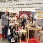 Na zdjęciu znajduje się grupa młodych ludzi, prawdopodobnie uczniów, stojących na czerwonym dywanie w hali wystawowej. Za nimi widoczne są banery promujące I LO im. Stanisława Staszica w Lublinie. Uczniowie mają na sobie białe koszulki z napisem "I ❤️ Staś", co jest skrótem od nazwy szkoły. Są to zarówno dziewczęta, jak i chłopcy, ubrani głównie w ciemne spodnie i sneakersy. Uczniowie stoją blisko siebie, część z nich trzyma broszury lub ulotki w dłoniach. W tle widoczny jest ekran telewizora, na którym wyświetla się obraz przedstawiający osobę stojącą na trawie z rozpostartymi ramionami. Banery z tyłu zawierają elementy graficzne, takie jak logo szkoły oraz wzory nawiązujące do nauki i technologii. Po prawej stronie znajduje się drewniany wysoki stół z broszurami i planszami edukacyjnymi, a obok niego stoi drewniany stołek barowy.