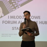 Na zdjęciu znajduje się młody mężczyzna stojący na tle ekranu z wyświetloną prezentacją. Mężczyzna ma jasną cerę, krótkie ciemne włosy i nosi okulary w cienkich, metalowych oprawkach. Ubrany jest w beżowy golf i ciemne spodnie. W lewej dłoni trzyma czarny notes lub tablet z kilkoma naklejkami, w tym jedną z postacią w kapeluszu i okularach przeciwsłonecznych. W prawej dłoni trzyma mikrofon, z którego korzysta podczas przemówienia. Jego postawa jest wyprostowana, wygląda na skupionego i zaangażowanego w wystąpienie. Tło stanowi duży ekran z projekcją slajdu prezentacji. Widnieje na nim tekst: "I MŁODZIEŻOWE FORUM AI CODE HUB STASZIC". W lewym górnym rogu slajdu znajduje się logo szkoły z wizerunkiem Stanisława Staszica oraz napisem „OGÓLNOKSZTAŁCĄCE IM. STANISŁAWA STASZICA W LUBLINIE”. W lewym dolnym rogu widoczny jest herb Lublina oraz tekst: "PREZYDENT MIASTA LUBLIN KRZYSZTOF ŻUK". Górna część slajdu ozdobiona jest grafiką przypominającą połączenia na płytce elektronicznej, co nawiązuje do tematyki nowoczesnych technologii i sztucznej inteligencji. Całość ma profesjonalny, minimalistyczny wygląd, z dominującymi odcieniami bieli, szarości i fioletu. Zdjęcie sugeruje, że wydarzenie odbywa się w formalnym lub edukacyjnym kontekście, prawdopodobnie w ramach forum poświęconego nowoczesnym technologiom i sztucznej inteligencji.