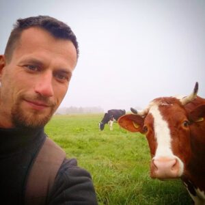 Mężczyzna znajduje się po lewej stronie zdjęcia, jego twarz zajmuje około jednej trzeciej obrazu. Ma krótkie, ciemne włosy i krótko przystrzyżoną brodę z wąsami. Ma jasną cerę, lekko uśmiechnięty wyraz twarzy i patrzy prosto w obiektyw. Ubrany jest w ciemną bluzę z wysokim kołnierzem i nosi brązowy plecak na jednym ramieniu, widoczny na pierwszym planie.
Tło i krajobraz:

W tle rozciąga się zielone pastwisko z trawiastym terenem, porośniętym niską, gęstą trawą. Widoczna jest lekka mgła lub zamglenie, które sprawia, że horyzont i oddalone drzewa są lekko rozmyte. Pogoda jest pochmurna – niebo jest jasnoszare, bez widocznego słońca.
Zwierzęta:

Najbardziej wyróżniającym się elementem zdjęcia obok mężczyzny jest krowa znajdująca się bardzo blisko obiektywu, po prawej stronie. To duża krowa w kolorze czerwono-brązowym z białą łatą biegnącą przez środek pyska, od czoła do nosa. Ma duże, jasne rogi zakrzywione ku górze i na boki oraz żółte kolczyki w uszach z widocznym numerem identyfikacyjnym. Krowa patrzy bezpośrednio w obiektyw i jest ustawiona przodem, jakby pozowała do zdjęcia razem z mężczyzną.

W dalszym planie widać drugą krowę – czarno-białą, która stoi i spokojnie skubie trawę. Znajduje się pośrodku kadru, w oddali.
Wrażenie ogólne:

Zdjęcie ma charakter pogodny i naturalny. Uchwycono moment bliskości człowieka i zwierzęcia w wiejskim, spokojnym krajobrazie. Obraz oddaje atmosferę spokoju, natury i codziennego życia na wsi.