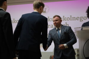 Mężczyzna w niebieskim garniturze o kraciastej fakturze i błękitnym krawacie podaje dłoń uczniowi w granatowym garniturze. Obaj patrzą na siebie z wyrazami uznania. W tle ekran z napisem oraz stos książek i kolorowe statuetki na stole.