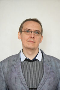 Na zdjęciu znajduje się mężczyzna w średnim wieku, sfotografowany od klatki piersiowej wzwyż, na jednolitym jasnym tle (najprawdopodobniej białym lub bardzo jasnokremowym). Zdjęcie jest portretowe, wykonane na wprost – postać patrzy nieco ponad obiektyw lub lekko w bok, z neutralnym, spokojnym wyrazem twarzy.
Wygląd osoby:

    Cera: jasna.

    Włosy: krótkie, w kolorze jasnobrązowym, przerzedzone na czubku głowy, ułożone w naturalny sposób, lekko sterczące.

    Twarz: owalna, bez zarostu.

    Oczy: nosi okulary z prostokątnymi szkłami i cienkimi, ciemnymi oprawkami.

    Wyraz twarzy: spokojny, poważny, z lekkim uniesieniem brwi – może sugerować skupienie lub zadumę.

Ubranie:

Mężczyzna jest ubrany elegancko, warstwowo:

    Marynarka: w szaro-fioletową kratę (klasyczny deseń), z szerokimi klapami i strukturalnymi ramionami.

    Sweter: ciemny, najprawdopodobniej czarny lub grafitowy, z pionowymi, cienkimi paskami w kontrastowym jasnym kolorze (np. białym lub szarym).

    Koszula: błękitna lub jasnoniebieska, z delikatnym wzorem lub fakturą. Kołnierzyk koszuli wystaje spod swetra, zapinany jest na guziki, a od środka podszyty ciemniejszym materiałem (możliwe, że granatowym).

Tło:

Jednolite, gładkie, bez ozdób ani elementów rozpraszających. Dzięki temu cała uwaga skupia się na postaci.

Wrażenie ogólne: Zdjęcie ma formalny charakter – może pochodzić z sesji portretowej pracownika naukowego, nauczyciela lub specjalisty w dziedzinie wymagającej oficjalnego wizerunku. Ubranie i postawa mężczyzny świadczą o profesjonalizmie i powadze.
