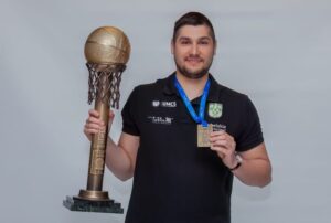 Na zdjęciu znajduje się młody mężczyzna o jasnej karnacji i krótkich, ciemnych włosach, stojący na neutralnym, jasnoszarym tle. Mężczyzna patrzy prosto w obiektyw i uśmiecha się delikatnie. Ubrany jest w czarną koszulkę polo z krótkim rękawem. Na koszulce znajdują się nadruki z logotypami instytucji i sponsorów. Po lewej stronie (z perspektywy widza) nadrukowane są logotypy: „UMCS” (Uniwersytet Marii Curie-Skłodowskiej), „Lublin AZS”, a po prawej stronie znajduje się herb AZS – zielono-biały tarczowy emblemat z literą „S” stylizowaną na kształt orła.

Mężczyzna trzyma dwa trofea:

    W lewej ręce – bardzo duży i efektowny puchar sportowy. Puchar składa się z wysokiego, cylindrycznego trzonu zakończonego kulą przypominającą złotą piłkę do koszykówki, oplecioną realistycznie odwzorowaną, brązową siatką do kosza. Siatka wykonana jest z metalowych ogniw, co dodaje pucharowi trójwymiarowego efektu. Całość osadzona jest na masywnej podstawie wykonanej z zielonego marmuru.

    W prawej ręce – mężczyzna trzyma złoty medal zawieszony na niebieskiej wstążce, którą ma przewieszoną przez szyję. Medal jest prostokątny i widnieje na nim napis w języku polskim: „MISTRZ POLSKI”. Symbolizuje zdobycie mistrzostwa Polski w dyscyplinie sportowej – prawdopodobnie koszykówce, sądząc po charakterze pucharu.

Postawa mężczyzny: Stoi wyprostowany i dumnie prezentuje trofea. Jego wyraz twarzy jest pogodny i zadowolony, co sugeruje, że jest dumny z osiągnięcia.

Tło zdjęcia: jednolite, gładkie, jasnoszare, bez dodatkowych elementów wizualnych, co pozwala skupić całą uwagę na postaci i trofeach.

Skojarzenia i kontekst: Zdjęcie prawdopodobnie zostało wykonane w studiu lub podczas oficjalnej sesji zdjęciowej po zdobyciu tytułu mistrza Polski przez drużynę sportową. Stroje, trofea oraz emblematy na koszulce wskazują na związek z uczelnią wyższą i sekcją koszykarską AZS UMCS Lublin.