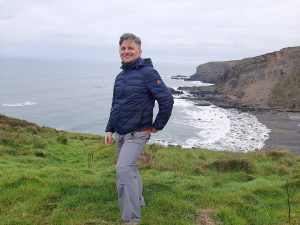 Na zdjęciu widoczny jest mężczyzna stojący na zielonym klifie z widokiem na skaliste wybrzeże i ocean. Zdjęcie zostało wykonane na zewnątrz, w chłodny, pochmurny dzień.
Opis osoby (na pierwszym planie, po lewej stronie zdjęcia):

    Mężczyzna w średnim wieku, o jasnej cerze i krótko ściętych, siwiejących włosach.

    Uśmiecha się szeroko i patrzy prosto w obiektyw. Jego postawa jest luźna, lekko skręcona w bok.

    Ubrany jest w ciemnoniebieską pikowaną kurtkę z kapturem i szare spodnie turystyczne z kieszeniami bocznymi. Z kurtki wystaje pomarańczowy fragment ubrania – prawdopodobnie koszulki.

    Stoi na zielonym, trawiastym wzniesieniu, z jedną ręką wsuniętą do kieszeni spodni, drugą swobodnie opuszczoną.

Tło (dominująca część krajobrazu):

    Ocean: rozciąga się po lewej stronie kadru aż po horyzont. Woda jest spokojna, stalowoszara, z delikatnymi falami rozbijającymi się o wybrzeże.

    Linia brzegowa: tworzy zatokę z wysokimi, stromymi klifami po prawej stronie zdjęcia. Klify są skaliste, brązowo-szare, z wyraźnymi warstwami geologicznymi.

    Plaża: poniżej klifów widać ciemną, kamienistą plażę, częściowo zalaną przez przypływ. Woda spływa po nierównym brzegu w postaci białych fal.

    Niebo: całkowicie zachmurzone, bez widocznego słońca, co daje jednolite, rozproszone światło bez cieni.

Wrażenie ogólne:

Zdjęcie ukazuje moment wypoczynku lub wędrówki w przybrzeżnym, surowym krajobrazie – prawdopodobnie w północnej Europie lub na Wyspach Brytyjskich. Mężczyzna wygląda na zrelaksowanego i szczęśliwego. Fotografia oddaje spokój, świeże powietrze i piękno dzikiej przyrody.