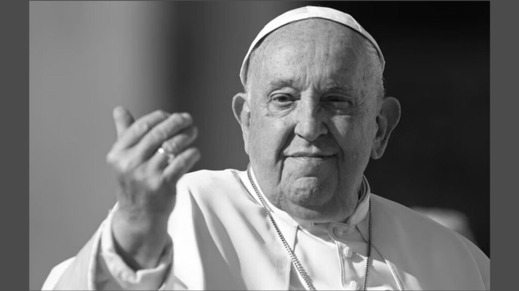 Zdjęcie czarno-białe, przedstawiające starszego mężczyznę w religijnym stroju katolickim, ujętego od klatki piersiowej w górę. Mężczyzna patrzy spokojnie przed siebie i lekko się uśmiecha. Jego twarz jest dobrze oświetlona naturalnym światłem dziennym. Ma okrągłą twarz, gładką skórę z widocznymi zmarszczkami, krótko ostrzyżone włosy (choć w większości łysiejące), a jego spojrzenie jest łagodne i spokojne. Na głowie nosi białą piuskę – tradycyjną, małą, okrągłą czapeczkę duchownych katolickich. Ubrany jest w białą sutannę (długi płaszcz duchowny sięgający do kostek, widoczny tutaj tylko do klatki piersiowej), z guzikami z przodu i ozdobnym, srebrnym łańcuszkiem z krzyżykiem, który delikatnie opada na jego pierś. Na serdecznym palcu prawej dłoni ma duży sygnet – pierścień, który często noszą papieże. Mężczyzna unosi prawą dłoń w geście błogosławieństwa lub pozdrowienia – dłoń jest lekko otwarta, palce rozchylone, skierowana ku przodowi i ku górze. Tło zdjęcia jest rozmyte i jednolite – prawdopodobnie przedstawia budynek lub ścianę w cieniu, co kieruje całą uwagę na postać. Zdjęcie emanuje spokojem, godnością i życzliwością. Styl ubrania i gest sugerują, że przedstawioną osobą jest prawdopodobnie papież w trakcie publicznego wystąpienia lub błogosławieństwa.