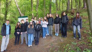 Zdjęcie przedstawia grupę około 17 młodych osób stojących na leśnej ścieżce w gęsto zalesionym terenie. Drzewa mają jasnozielone liście – typowe dla wiosny. W tle widoczna jest tablica informacyjna z napisem „Nadleśnictwo Kołczygłowy” oraz opisem przyrodniczym. Uczniowie są ubrani w ciemne, sportowe i turystyczne ubrania – kurtki, bluzy, czapki, część z nich ma plecaki. Wszyscy stoją blisko siebie i pozują do zdjęcia, część się uśmiecha. Tło to gęsty las z prostymi, wysokimi drzewami, na ziemi ściółka leśna.