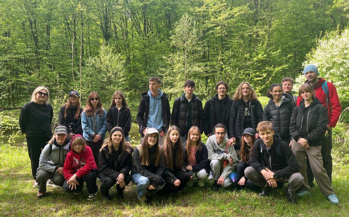 Zdjęcie grupowe wykonane na skraju polany. Przed aparat ustawiła się cała grupa – około 24 osób, część w kucki, część stoi. Z tyłu za nimi gęsty las liściasty, z bardzo jasnozielonymi liśćmi. Po lewej stronie nauczycielka w czarnym ubraniu i okularach przeciwsłonecznych. Obok niej uczniowie – część ubrana w czapki z daszkiem, kurtki, dresy. Po prawej stronie mężczyzna w czerwonej kurtce i niebieskiej czapce. Wszyscy pozują i patrzą w stronę aparatu. Na pierwszym planie zielona trawa, widoczne też pojedyncze gałęzie i patyki.