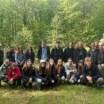 Zdjęcie grupowe wykonane na skraju polany. Przed aparat ustawiła się cała grupa – około 24 osób, część w kucki, część stoi. Z tyłu za nimi gęsty las liściasty, z bardzo jasnozielonymi liśćmi. Po lewej stronie nauczycielka w czarnym ubraniu i okularach przeciwsłonecznych. Obok niej uczniowie – część ubrana w czapki z daszkiem, kurtki, dresy. Po prawej stronie mężczyzna w czerwonej kurtce i niebieskiej czapce. Wszyscy pozują i patrzą w stronę aparatu. Na pierwszym planie zielona trawa, widoczne też pojedyncze gałęzie i patyki.
