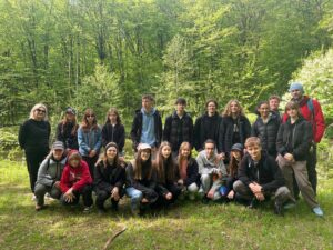 Zdjęcie grupowe wykonane na skraju polany. Przed aparat ustawiła się cała grupa – około 24 osób, część w kucki, część stoi. Z tyłu za nimi gęsty las liściasty, z bardzo jasnozielonymi liśćmi. Po lewej stronie nauczycielka w czarnym ubraniu i okularach przeciwsłonecznych. Obok niej uczniowie – część ubrana w czapki z daszkiem, kurtki, dresy. Po prawej stronie mężczyzna w czerwonej kurtce i niebieskiej czapce. Wszyscy pozują i patrzą w stronę aparatu. Na pierwszym planie zielona trawa, widoczne też pojedyncze gałęzie i patyki.
