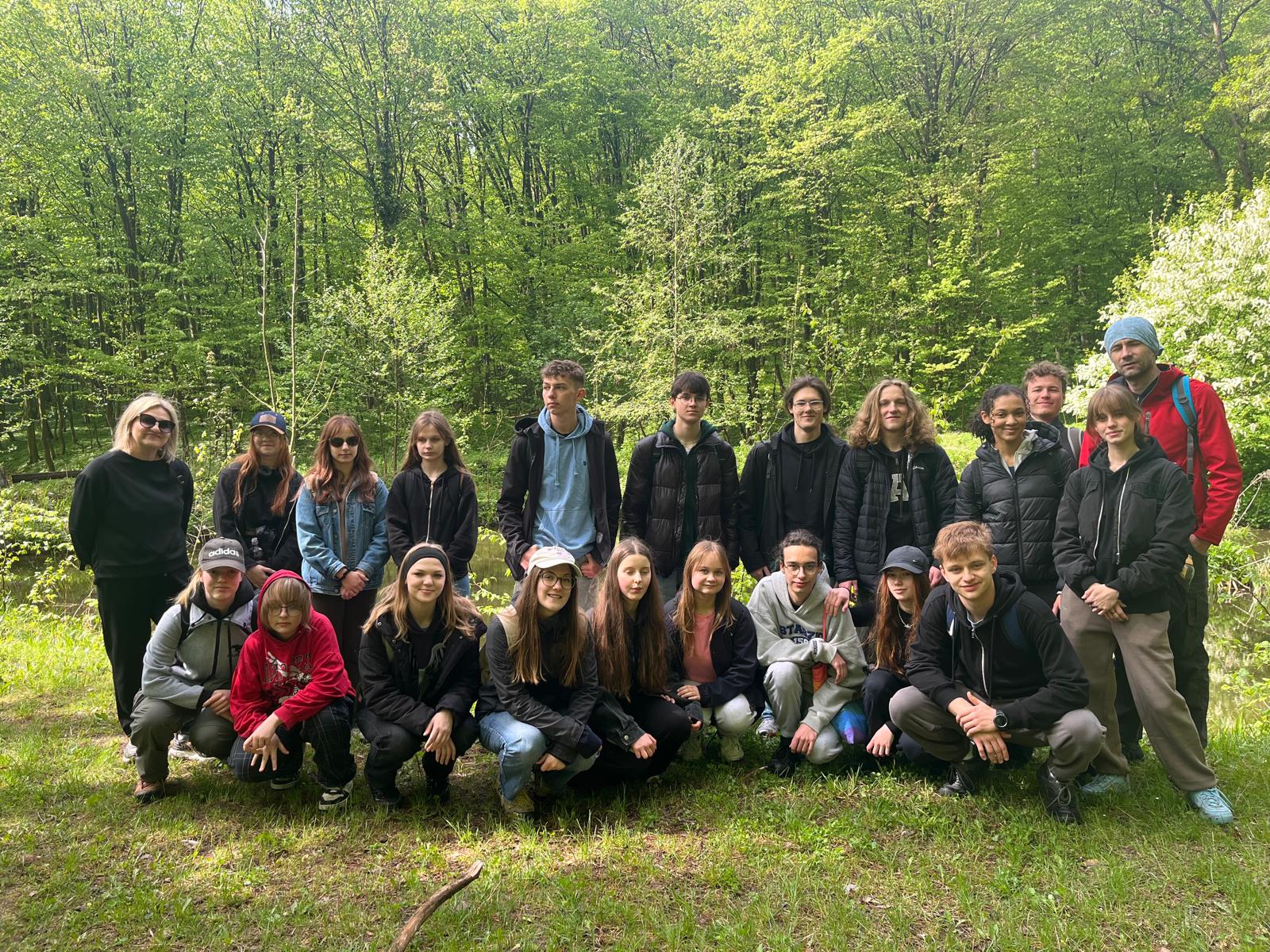 Zdjęcie grupowe wykonane na skraju polany. Przed aparat ustawiła się cała grupa – około 24 osób, część w kucki, część stoi. Z tyłu za nimi gęsty las liściasty, z bardzo jasnozielonymi liśćmi. Po lewej stronie nauczycielka w czarnym ubraniu i okularach przeciwsłonecznych. Obok niej uczniowie – część ubrana w czapki z daszkiem, kurtki, dresy. Po prawej stronie mężczyzna w czerwonej kurtce i niebieskiej czapce. Wszyscy pozują i patrzą w stronę aparatu. Na pierwszym planie zielona trawa, widoczne też pojedyncze gałęzie i patyki.