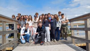 Na zdjęciu widoczna jest grupa około 20 uczniów w wieku 16–19 lat, siedzących i stojących na drewnianej kładce przecinającej torfowisko. Uczestnicy ubrani są w luźne, sportowe stroje: bluzy, t-shirty, dresy, jeansy, sportowe buty. Niektórzy mają na głowach czapki z daszkiem, a kilku z nich nosi plecaki.

Tło zdjęcia stanowi naturalne torfowisko porośnięte niskimi drzewami iglastymi, głównie sosnami o cienkich pniach i delikatnych gałązkach. Wśród traw i mchów widoczne są białe, puszyste kwiaty wełnianek, które rosną licznie, tworząc efekt białych kropek na tle zieleni.

Uczniowie siedzą lub kucają w rzędzie, blisko siebie, tworząc półokrąg lub luźną linię, część osób patrzy w stronę fotografa i uśmiecha się. Wyrazy twarzy wyrażają zadowolenie i radość.