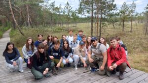 Grupa młodzieży siedzi na trawie na skraju torfowiska, odpoczywając po spacerze. Uczniowie siedzą w kręgu, niektórzy jedzą przekąski, inni rozmawiają lub przeglądają zdjęcia na telefonach.

W tle widoczne są wysokie trawy, pojedyncze drzewa i fragmenty torfowiska z białymi kwiatami wełnianek. Atmosfera jest spokojna i relaksująca.