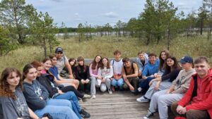 Uczestnicy to młodzi ludzie, prawdopodobnie uczniowie szkoły średniej, w wieku około 16–19 lat. Większość ubrana jest w luźne, sportowe stroje: bluzy, t-shirty, dresy, jeansy, sportowe buty. Kilka osób ma na głowach czapki z daszkiem. Niektórzy mają plecaki.
Tło zdjęć:

    Młodzież siedzi lub klęczy na drewnianej kładce w środku terenu podmokłego, z drewnianą barierką za nimi.

    Teren dookoła to naturalne torfowisko porośnięte niskimi drzewami iglastymi, głównie sosnami o cienkich pniach i delikatnych gałązkach. Wśród traw i mchów widoczne są białe, puszyste kwiaty wełnianek, które rosną licznie, tworząc efekt białych kropek na tle zieleni.

    W oddali za grupą widać jezioro lub rozlewisko, z którego wystają fragmenty traw i niskiej roślinności. Niebo jest jasne, z lekkim zachmurzeniem, sugerując spokojny, letni lub wiosenny dzień.

Szczegółowy opis grupy:

    Młodzież siedzi lub kuca w rzędzie, blisko siebie, tworząc półokrąg lub luźną linię, część osób patrzy w stronę fotografa i uśmiecha się.

    Wyrazy twarzy: zadowolenie, uśmiechy, czasem neutralne miny.

    Kolorystyka ubrań: przeważają szarości, czernie, granaty, błękity, biele. Kilka osób ma na sobie ubrania w żywszych kolorach, np. różowe bluzy, czerwoną kurtkę.

    Wśród uczestników jest kilka osób z charakterystycznymi detalami:

        Młody mężczyzna w czerwonej kurtce i spodniach moro, siedzący blisko przedniego rzędu.

        Dziewczyna w szarym swetrze z napisem „Soft Road Lublin”.

        Chłopak w beżowej bluzie z grafiką i czapce z daszkiem, trzymający ręce na kolanach.

        Kilka osób ma splecione warkocze, długie rozpuszczone włosy lub spięte w kucyki.

Dodatkowe detale dostępnościowe:

    Drewno pomostu jest jasne, częściowo wypłowiałe od słońca, z wyraźnymi słojami i strukturą drewna. Na pomostach widoczne są metalowe śruby lub elementy mocujące.

    Na zdjęciach nie ma przeszkód terenowych ani elementów nieprzyjaznych – przestrzeń jest równa, kładka umożliwia swobodne poruszanie się osobom z ograniczoną mobilnością.

    W tle nie widać intensywnej infrastruktury – to naturalny teren z ograniczoną ingerencją człowieka, idealny do zajęć edukacyjnych.