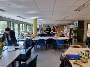 Zdjęcie wykonane w jasnym, przestronnym pomieszczeniu przypominającym pokój nauczycielski lub pracownię szkolną. W centralnej części sali znajdują się stoły ustawione w prostokąt, otoczone krzesłami, niektóre z nich są niebieskie, inne czarne. Na pierwszym planie po lewej stronie kobieta z długimi brązowymi włosami pochyla się nad papierami na biurku, ubrana w ciemną kurtkę i okulary. Na stołach widoczne są różne przedmioty: tulipany w wazonie, jedzenie (m.in. banany, jabłka), napoje, przybory piśmiennicze, kartki i segregatory. W głębi sali, przy ścianie, stoi grupa około siedmiu osób – młodych ludzi i nauczycieli – trzymających kartki i rozmawiających ze sobą. Ściany z lewej strony są przeszklone, przez okna widać zieleń drzew. Na ścianach i szafkach widoczne są segregatory, pudełka i różne wyposażenie biurowe. Atmosfera nieformalna, skupiona i życzliwa.