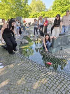 Na zdjęciu grupa nastolatków, głównie dziewcząt, zgromadzona jest przy fontannie w przestrzeni miejskiej. Fontanna ma formę niskiej, szerokiej rynny z płytkim strumieniem wody, płynącej przez nieregularnie ułożone kamienie i mozaikę z kostki brukowej. Woda jest czysta, widać w niej odbicia postaci i drzew. Dwie dziewczyny na pierwszym planie klęczą, a jedna z nich wypuszcza na wodę plastikową butelkę z czerwonymi dekoracjami, co sugeruje, że odbywa się zabawa polegająca na puszczaniu butelek po wodzie. Pozostałe osoby, około 15, stoją wokół i przyglądają się z zainteresowaniem, niektóre się śmieją. Tło wypełniają zielone drzewa i jasne światło słoneczne.