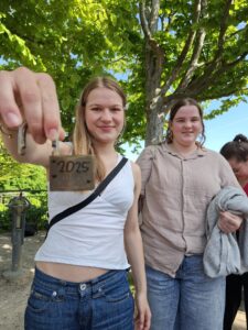 Na zdjęciu centralną postacią jest młoda dziewczyna z jasnymi włosami, która zbliża do obiektywu kłódkę w kolorze miedzianym. Na kłódce wygrawerowany jest napis „2025 ALLES ♥”. Dziewczyna uśmiecha się lekko, ubrana jest w biały top na ramiączkach i dżinsy, przez ramię ma przewieszoną czarną torebkę z paskiem. Obok niej stoi inna dziewczyna w beżowej koszuli i jasnych jeansach, trzymająca sweter w ręku. Obie stoją na tle jasnej, piaszczystej ścieżki w parku, a za nimi widać duże zielone drzewa z gęstym listowiem. Niebo jest bezchmurne, a światło słoneczne rozjaśnia całe zdjęcie.