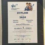 Obraz przedstawia dyplom oprawiony w czarną ramkę z metalicznym połyskiem, umieszczony pionowo. Tło samego dyplomu jest białe, tekst wydrukowany w kolorze czarnym i granatowym, z kolorowymi elementami graficznymi u góry. Szczegółowy opis graficzny i tekstowy: Górna część dyplomu: Po lewej stronie znajduje się logo SIGG 23, składające się z dużych liter „SIGG” (pierwsze litery nazwy „Szkolna Internetowa Gra Giełdowa”), napisane ciemnoniebieskim kolorem, z nieco pochyloną do przodu literą „G”. Nad drugim „G” widnieje liczba „23”, oznaczająca edycję konkursu. Pod logotypem znajduje się pełna nazwa: „Szkolna Internetowa Gra Giełdowa”. Po prawej stronie, w górnym narożniku, znajduje się grafika dwojga młodych ludzi: Po lewej stoi chłopiec z brązowymi włosami ułożonymi do góry, w niebieskiej bluzie z kapturem, założonymi rękami i lekkim uśmiechem. Obok niego stoi dziewczyna z ciemnymi włosami upiętymi w niedbały kok, ubrana w brązową kurtkę i niebieski szalik. Ona także jest uśmiechnięta. Oboje są pokazani od pasa w górę, patrzą z lekkim optymizmem, w tle za nimi biegnie niebieski wykres giełdowy, który unosi się ku górze. Środkowa część dyplomu: Na środku, dużymi, pogrubionymi literami widnieje tytuł: DYPLOM dla zespołu 3A24 w składzie: Bolesław Główczyk Maksymilian Motyka pod opieką Małgorzaty Mańkowskiej z I Liceum Ogólnokształcącego im. Stanisława Staszica w Lublinie Dolna część dyplomu: Tekst poniżej mówi: za zajęcie 4. miejsca w finale XXIII edycji Szkolnej Internetowej Gry Giełdowej organizowanej przez Giełdę Papierów Wartościowych w Warszawie oraz Fundację GPW Warszawa, 29 kwietnia 2025 r. Poniżej znajdują się trzy podpisy (wydrukowane z nazwiskami): Tomasz Bardziłowski – Prezes Zarządu Giełdy Papierów Wartościowych w Warszawie dwa inne podpisy (nieczytelne na zdjęciu, ale obecne w dolnej części dokumentu) Dolny margines dyplomu: Znajdują się logotypy organizatorów i partnerów: GPW (Giełda Papierów Wartościowych) Fundacja GPW PAGA (Fundacja im. Lesława A. Pagi) inne instytucje partnerskie (logotypy nieczytelne, ale obecne)