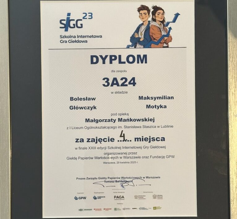Obraz przedstawia dyplom oprawiony w czarną ramkę z metalicznym połyskiem, umieszczony pionowo. Tło samego dyplomu jest białe, tekst wydrukowany w kolorze czarnym i granatowym, z kolorowymi elementami graficznymi u góry. Szczegółowy opis graficzny i tekstowy: Górna część dyplomu: Po lewej stronie znajduje się logo SIGG 23, składające się z dużych liter „SIGG” (pierwsze litery nazwy „Szkolna Internetowa Gra Giełdowa”), napisane ciemnoniebieskim kolorem, z nieco pochyloną do przodu literą „G”. Nad drugim „G” widnieje liczba „23”, oznaczająca edycję konkursu. Pod logotypem znajduje się pełna nazwa: „Szkolna Internetowa Gra Giełdowa”. Po prawej stronie, w górnym narożniku, znajduje się grafika dwojga młodych ludzi: Po lewej stoi chłopiec z brązowymi włosami ułożonymi do góry, w niebieskiej bluzie z kapturem, założonymi rękami i lekkim uśmiechem. Obok niego stoi dziewczyna z ciemnymi włosami upiętymi w niedbały kok, ubrana w brązową kurtkę i niebieski szalik. Ona także jest uśmiechnięta. Oboje są pokazani od pasa w górę, patrzą z lekkim optymizmem, w tle za nimi biegnie niebieski wykres giełdowy, który unosi się ku górze. Środkowa część dyplomu: Na środku, dużymi, pogrubionymi literami widnieje tytuł: DYPLOM dla zespołu 3A24 w składzie: Bolesław Główczyk Maksymilian Motyka pod opieką Małgorzaty Mańkowskiej z I Liceum Ogólnokształcącego im. Stanisława Staszica w Lublinie Dolna część dyplomu: Tekst poniżej mówi: za zajęcie 4. miejsca w finale XXIII edycji Szkolnej Internetowej Gry Giełdowej organizowanej przez Giełdę Papierów Wartościowych w Warszawie oraz Fundację GPW Warszawa, 29 kwietnia 2025 r. Poniżej znajdują się trzy podpisy (wydrukowane z nazwiskami): Tomasz Bardziłowski – Prezes Zarządu Giełdy Papierów Wartościowych w Warszawie dwa inne podpisy (nieczytelne na zdjęciu, ale obecne w dolnej części dokumentu) Dolny margines dyplomu: Znajdują się logotypy organizatorów i partnerów: GPW (Giełda Papierów Wartościowych) Fundacja GPW PAGA (Fundacja im. Lesława A. Pagi) inne instytucje partnerskie (logotypy nieczytelne, ale obecne)