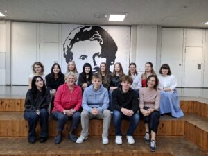 Grupa czternastu osób, w tym dwie nauczycielki i dwanaścioro uczniów, pozuje do zdjęcia na drewnianych stopniach w przestrzeni szkolnej. W tle biała ściana z czarnym, dużym graficznym portretem Fryderyka Chopina (lub innej znanej osoby). Osoby są uśmiechnięte, pozują w dwóch rzędach: siedzącym i stojącym. Wszyscy są ubrani schludnie, w codzienne ubrania.