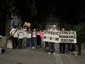 Tło i otoczenie:
Zdjęcie zostało wykonane nocą, na zewnątrz przed budynkiem (najpewniej szkolnym), który jest częściowo oświetlony światłem latarni oraz światłem z wnętrza. W tle widać drzewa iglaste oraz wejście do budynku z kolumnami i schodami.
Osoby:
Na zdjęciu stoi około 14 osób – większość młodzież, a także kilka dorosłych. Wszyscy pozują razem, tworząc zwartą grupę. Są uśmiechnięci i w dobrych nastrojach.
Ubrania:
Większość ma na sobie białe koszulki z bordowym nadrukiem „STASZIC RUNNING TEAM”. Dwie osoby pośrodku wyróżniają się: jedna kobieta ma różową kurtkę, druga – ciemnoróżową koszulkę z tym samym napisem. Jedna osoba trzyma pomarańczowy transparent z napisem „GO GORYL”.
Transparenty:
Po prawej stronie grupy dwie osoby trzymają duże, ręcznie wykonane transparenty z hasłami:
Lewy transparent: „STASZIC R U N !” – każda litera napisana drukowanymi czarnymi literami, w słowie RUN litery są ozdobione rysunkami stylizowanych biegaczy.
Prawy transparent: „STASZIC RUNNING DREAM” – także ręcznie wykonany, litery są duże, czarne z czerwonym obrysem.