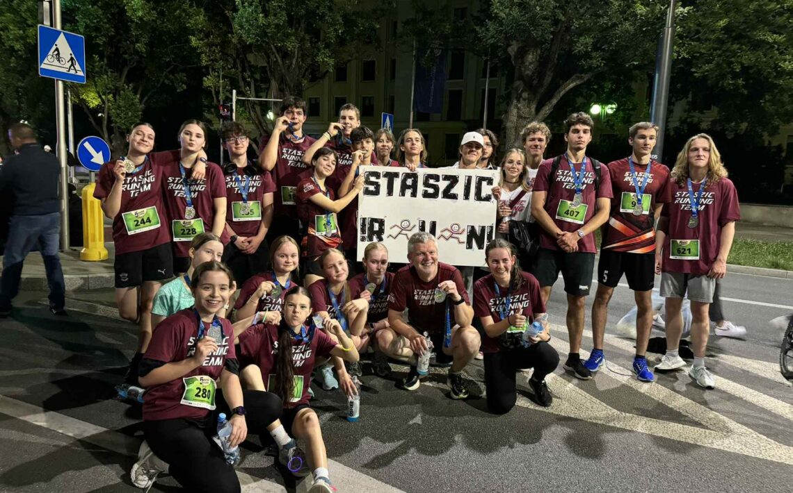 Tło i otoczenie: Zdjęcie wykonane nocą na ulicy, w pobliżu przejścia dla pieszych. Widoczna jest sygnalizacja świetlna, drzewa oraz oświetlone budynki w tle. Widać oznaczenia drogowe – znak przejścia dla pieszych i ścieżki rowerowej. Teren jest dobrze oświetlony latarniami. Osoby: Na zdjęciu znajduje się duża grupa – około 25 osób. Wszyscy pozują razem po zakończonym biegu. Część stoi, część kuca na pierwszym planie. Wszyscy są radośni, część pokazuje medale lub butelki z wodą. Ubrania i identyfikacja: Wszyscy mają na sobie bordowe koszulki z napisem „STASZIC RUNNING TEAM” w białym kolorze. Większość z nich ma przypięte na brzuchu zielone lub niebieskie numery startowe. Numery startowe to m.in. 244, 228, 280, 407, 621. Widoczne są też buty sportowe i opaski. Transparent: W centrum zdjęcia znajduje się ten sam biały transparent, co na poprzednim zdjęciu, z napisem „STASZIC R U N!” – litery ozdobione rysunkami biegnących postaci.