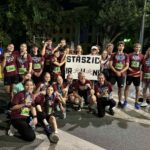 Tło i otoczenie: Zdjęcie wykonane nocą na ulicy, w pobliżu przejścia dla pieszych. Widoczna jest sygnalizacja świetlna, drzewa oraz oświetlone budynki w tle. Widać oznaczenia drogowe – znak przejścia dla pieszych i ścieżki rowerowej. Teren jest dobrze oświetlony latarniami. Osoby: Na zdjęciu znajduje się duża grupa – około 25 osób. Wszyscy pozują razem po zakończonym biegu. Część stoi, część kuca na pierwszym planie. Wszyscy są radośni, część pokazuje medale lub butelki z wodą. Ubrania i identyfikacja: Wszyscy mają na sobie bordowe koszulki z napisem „STASZIC RUNNING TEAM” w białym kolorze. Większość z nich ma przypięte na brzuchu zielone lub niebieskie numery startowe. Numery startowe to m.in. 244, 228, 280, 407, 621. Widoczne są też buty sportowe i opaski. Transparent: W centrum zdjęcia znajduje się ten sam biały transparent, co na poprzednim zdjęciu, z napisem „STASZIC R U N!” – litery ozdobione rysunkami biegnących postaci.