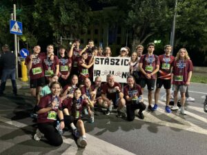 Tło i otoczenie:
Zdjęcie wykonane nocą na ulicy, w pobliżu przejścia dla pieszych. Widoczna jest sygnalizacja świetlna, drzewa oraz oświetlone budynki w tle. Widać oznaczenia drogowe – znak przejścia dla pieszych i ścieżki rowerowej. Teren jest dobrze oświetlony latarniami.
Osoby:
Na zdjęciu znajduje się duża grupa – około 25 osób. Wszyscy pozują razem po zakończonym biegu. Część stoi, część kuca na pierwszym planie. Wszyscy są radośni, część pokazuje medale lub butelki z wodą.
Ubrania i identyfikacja:
Wszyscy mają na sobie bordowe koszulki z napisem „STASZIC RUNNING TEAM” w białym kolorze. Większość z nich ma przypięte na brzuchu zielone lub niebieskie numery startowe. Numery startowe to m.in. 244, 228, 280, 407, 621. Widoczne są też buty sportowe i opaski.
Transparent:
W centrum zdjęcia znajduje się ten sam biały transparent, co na poprzednim zdjęciu, z napisem „STASZIC R U N!” – litery ozdobione rysunkami biegnących postaci.