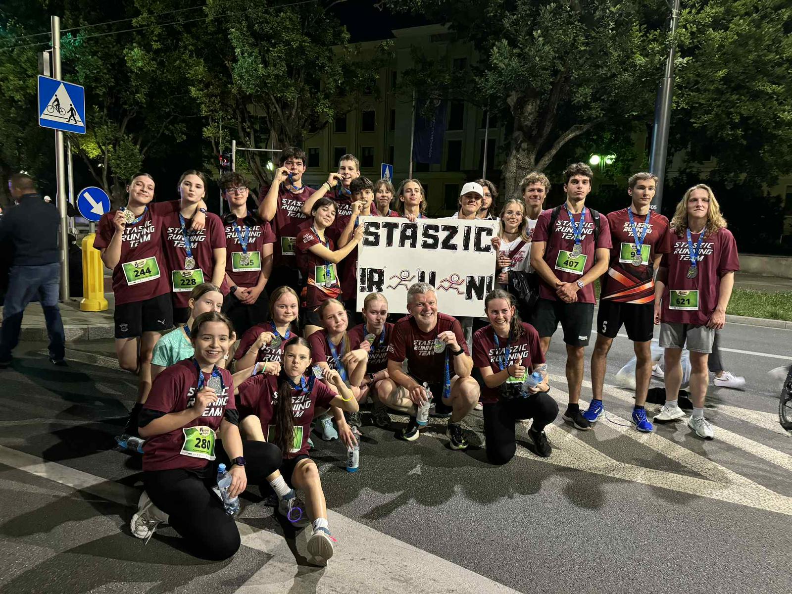 Tło i otoczenie: Zdjęcie wykonane nocą na ulicy, w pobliżu przejścia dla pieszych. Widoczna jest sygnalizacja świetlna, drzewa oraz oświetlone budynki w tle. Widać oznaczenia drogowe – znak przejścia dla pieszych i ścieżki rowerowej. Teren jest dobrze oświetlony latarniami. Osoby: Na zdjęciu znajduje się duża grupa – około 25 osób. Wszyscy pozują razem po zakończonym biegu. Część stoi, część kuca na pierwszym planie. Wszyscy są radośni, część pokazuje medale lub butelki z wodą. Ubrania i identyfikacja: Wszyscy mają na sobie bordowe koszulki z napisem „STASZIC RUNNING TEAM” w białym kolorze. Większość z nich ma przypięte na brzuchu zielone lub niebieskie numery startowe. Numery startowe to m.in. 244, 228, 280, 407, 621. Widoczne są też buty sportowe i opaski. Transparent: W centrum zdjęcia znajduje się ten sam biały transparent, co na poprzednim zdjęciu, z napisem „STASZIC R U N!” – litery ozdobione rysunkami biegnących postaci.