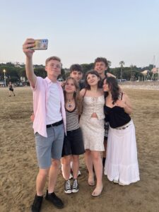 Sześć młodych osób robi wspólne zdjęcie selfie na piaszczystej plaży. Osoba z przodu po lewej stronie (prawdopodobnie chłopak) trzyma telefon z kolorowym etui i wykonuje zdjęcie. Ma na sobie różową koszulę, jasne spodenki i okulary. Pozostali pozują w różnych pozach, uśmiechnięci, w letnich stylizacjach – sukienki, topy, krótkie spodenki. W tle widać piasek, niskie drzewa i latarnie. Uczniowie sprawiają wrażenie radosnych i zrelaksowanych.