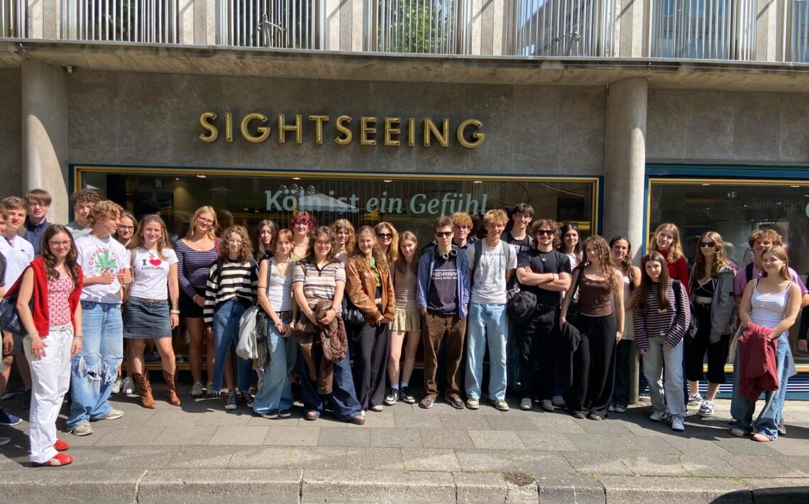 Zdjęcie grupowe zrobione na zewnątrz, przed witryną sklepową lub punktem informacyjnym z dużym napisem „SIGHTSEEING” nad głowami uczestników. Przez szybę widoczny napis: „Köln ist ein Gefühl” (Kolonia to uczucie). Przed budynkiem stoi grupa około 30 młodych osób, uśmiechniętych, ubranych swobodnie i letnio. Ulica jest brukowana, dzień jest słoneczny.