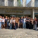 Zdjęcie grupowe zrobione na zewnątrz, przed witryną sklepową lub punktem informacyjnym z dużym napisem „SIGHTSEEING” nad głowami uczestników. Przez szybę widoczny napis: „Köln ist ein Gefühl” (Kolonia to uczucie). Przed budynkiem stoi grupa około 30 młodych osób, uśmiechniętych, ubranych swobodnie i letnio. Ulica jest brukowana, dzień jest słoneczny.