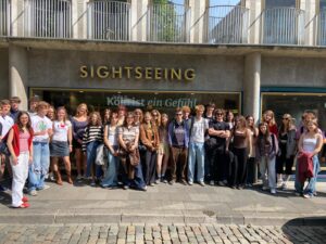 Zdjęcie grupowe zrobione na zewnątrz, przed witryną sklepową lub punktem informacyjnym z dużym napisem „SIGHTSEEING” nad głowami uczestników. Przez szybę widoczny napis: „Köln ist ein Gefühl” (Kolonia to uczucie). Przed budynkiem stoi grupa około 30 młodych osób, uśmiechniętych, ubranych swobodnie i letnio. Ulica jest brukowana, dzień jest słoneczny.
