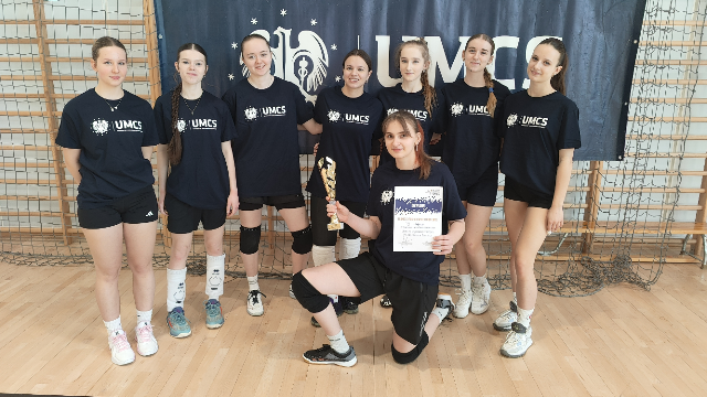 Grupa siatkarek z UMCS: Tło: Drewniana podłoga hali sportowej, ściana z siatką zabezpieczającą, na ścianie duży ciemnoniebieski baner z białym napisem „UMCS” i logo uczelni – herb przedstawiający sylwetkę sowy oraz symbol ognia i gwiazd. Uczestniczki: Osiem młodych kobiet ubranych w granatowe koszulki sportowe z logo „UMCS” na piersi, czarne lub białe spodenki sportowe, niektóre z ochraniaczami na kolana (czarne lub białe), białe lub kolorowe buty sportowe. Pierwszy plan: Jedna z dziewczyn klęczy z przodu na jedno kolano, trzyma w jednej ręce puchar złoty, a w drugiej dyplom – biała kartka formatu A4 z ciemnoniebieskim tekstem i logo uczelni. Reszta drużyny: Stoi z tyłu w półokręgu, uśmiechnięte, patrzą w stronę aparatu. Atmosfera: Radość z wygranej, zadowolenie z sukcesu sportowego, drużyna w pełnym składzie.
