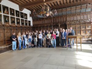 Grupowe zdjęcie wykonane w zabytkowym wnętrzu, prawdopodobnie w muzeum lub ratuszu. Uczestnicy stoją pod rzeźbioną, drewnianą boazerią, nad którą zawieszono kilkanaście portretów w złotych ramach (prawdopodobnie postaci historyczne). Nad grupą wisi ozdobny żyrandol z herbami i świecami. Wnętrze jest majestatyczne i bogato zdobione. Grupa liczy około 30 osób – zarówno młodzież, jak i kilku dorosłych opiekunów.