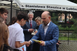 Na pierwszym planie młody chłopak w białej koszuli z krótkim rękawem odbiera książkę i ściska dłoń mężczyzny w niebieskiej marynarce w kratę. Mężczyzna ma długą brodę, łysinę na czubku głowy i długie włosy z tyłu głowy. W tle widać kilka osób, w tym starszego mężczyznę w garniturze i kobietę z długimi blond włosami podpisującą książkę. Namiot ma nadrukowany napis: „im. Stanisława Staszica w Lublinie”.
