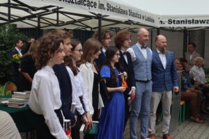 Grupa uczniów (8 osób) pozuje do zdjęcia pamiątkowego razem z dwoma nauczycielami – jednym w błękitnej marynarce w kratę i drugim w jasnoszarym garniturze w kratę z muszką. Uczniowie trzymają książki. Wszyscy są odświętnie ubrani – białe koszule, sukienki, garnitury. Tło stanowi biały namiot z napisem „im. Stanisława Staszica w Lublinie”.