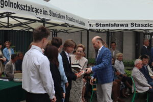 Grupa uczniów odbiera nagrody od mężczyzny w niebieskiej marynarce. Jeden z uczniów z blond włosami odbiera książkę, stojąc obok kobiety w beżowym stroju, która także trzyma książki. W tle – osoby siedzące na krzesłach, zielone krzesła szkolne, część osób z książkami. Namiot z napisem szkoły widoczny u góry.