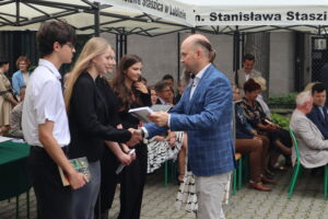 Trójka uczniów, w tym jedna dziewczyna z blond włosami w czarnej marynarce, odbiera książki od mężczyzny w niebieskiej marynarce w kratę. Dziewczyna ściska jego dłoń. Jeden z chłopców trzyma książkę Stephena Kinga. W tle widoczni goście siedzący pod namiotem.