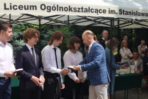 Kilku uczniów stoi w kolejce po odbiór książek. Mężczyzna w niebieskiej marynarce wręcza książkę chłopakowi w białej koszuli. Obok niego stoi dziewczyna w białej bluzce z falbankami i czarnej, plisowanej spódnicy, trzymająca książkę. Za nimi – nauczyciele i stosy książek na stole.