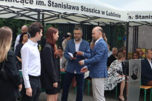 Troje uczniów rozmawia z dwoma mężczyznami – jeden z nich rozdaje książki, drugi notuje coś w zeszycie. Jedna z uczennic ma rude, długie włosy i czarny strój. W tle widoczna publiczność siedząca pod namiotem.