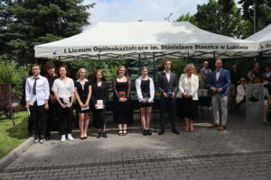 Dziewięcioro uczniów (5 dziewczyn, 4 chłopców) pozuje do zdjęcia z książkami w dłoniach. Stoją przed namiotem z napisem „I Liceum Ogólnokształcące im. Stanisława Staszica w Lublinie”. Obok nich stoją nauczyciele. Tło stanowi zieleń, namioty i zgromadzeni goście.