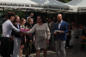 Ujęcie przedstawia kobietę w kremowej sukience, z ciemnymi włosami i okularami, ściskającą dłoń młodego chłopaka w białej koszuli. W tle stoi grupa uczennic z kwiatami, ubranych odświętnie – m.in. w ciemne marynarki i sukienki. Po prawej stronie mężczyzna z długą brodą w niebieskiej marynarce trzyma stos książek. Scena odbywa się na zewnątrz pod namiotem z napisem „im. Stanisława Staszica w Lublinie”.