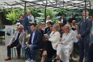 Publiczność siedząca pod namiotem – widoczne są dwie pierwsze ławki z nauczycielami i zaproszonymi gośćmi. Kobiety w jasnych ubraniach, eleganckie fryzury, mężczyźni w garniturach. W tle osoby stojące – jedna kobieta trzyma bukiet słoneczników. Po lewej stronie mównica z logo szkoły – czarny profil Stanisława Staszica na tle białym.