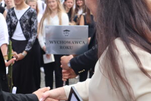 Zbliżenie na wręczenie nagrody „Wychowawca Roku”. Tabliczka z napisem „Wychowawca Roku” trzymana przez uczennicę w czarnym stroju. Obok kobieta w kremowej sukience z długimi, brązowymi włosami ściska rękę nagradzającego. W tle uczniowie stojący w szeregu – ubrane głównie w białe i czarne stroje galowe.
